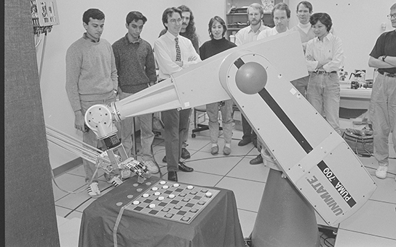 A black and white photo of several people standing in a lab watching a robot arm play checkers.