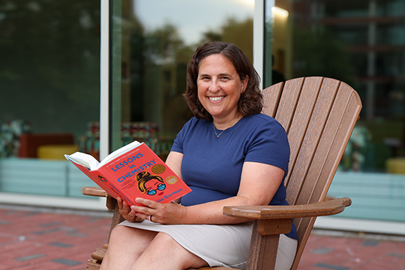 Wendi Heinzelman sits in an Adirondack chair in front of Wegmans Hall with the book "Lessons in Chemistry" in her hands.