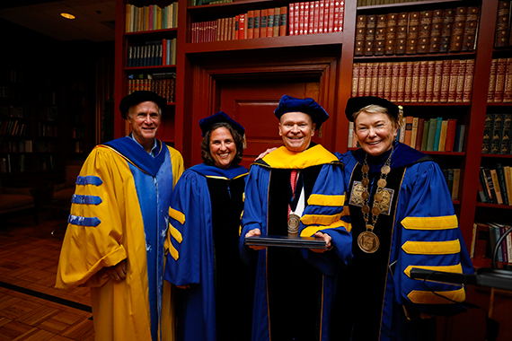 Tom Brown, Wendi Heinzelman, John Bruning, and Sarah Mangelsdorft wearing blue and gold academic regalia while smiling in a library.