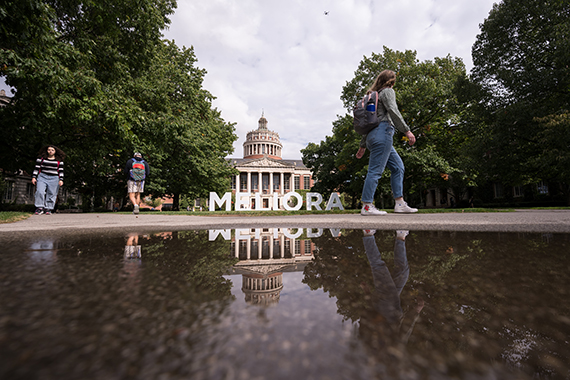 A low profile view of the Eastman Quadrangle with the large letters spelling "MELIORA" and the Rush Rhees Library reflecting in a puddle.