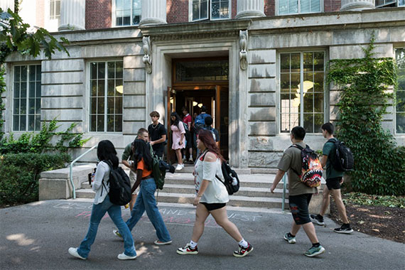 Students walk in and out of a classical academic building entrance with large windows and columns.