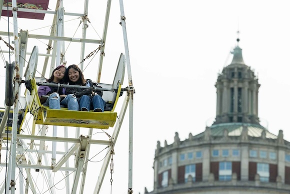 two students ride a ferris wheel with Rush Rhees Library tower in the background.