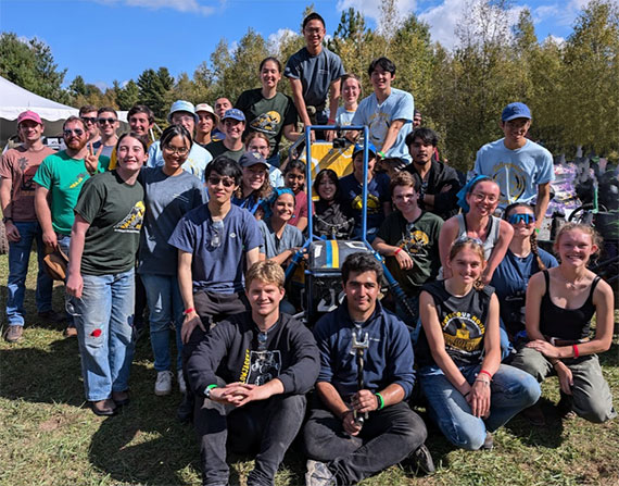 Dozens of Rochester students gather around the Baja SAE car outdoors.