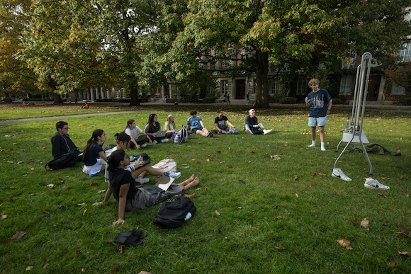 Students sit in a semicircle on the grass facing another student who is instructing them