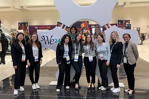 Ten women engineering students pose in front of a 10-foot gear with the WE24 logo on it.