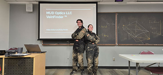 Two optics students pose back to back in the front of the classroom while wearing mud-covered clothes.