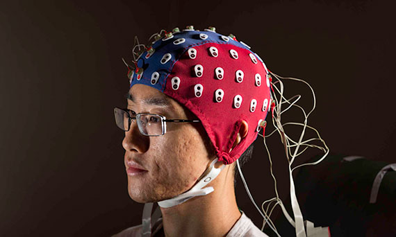 A person with glasses wears a blue and red EEG cap with wires coming out the back.