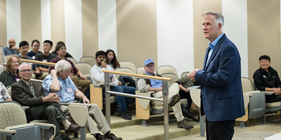 Tom Brown speaks in front of an auditorium in Goergen Hall crowded with students and faculty.