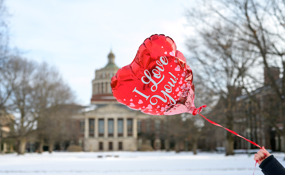 A hand holds a red balloon with the words I Love You. Rush Rhees Library is in the background. 