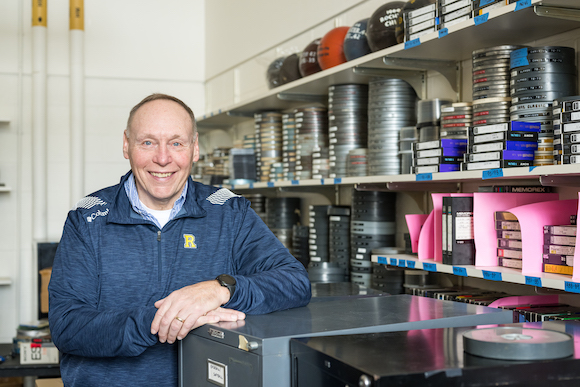 Terry Gurnett in blue Rochester jacket. He is pictured in front of shelves of film canisters. 
