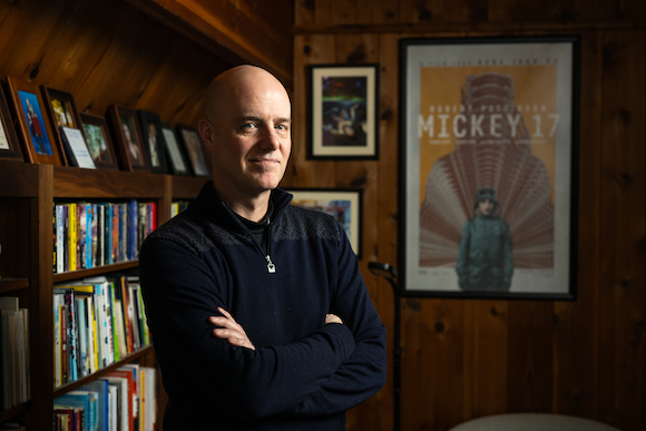 Edward Ashton ‘95 PhD is photographed at his home. A Mickey 17 film poster hangs behind him. 