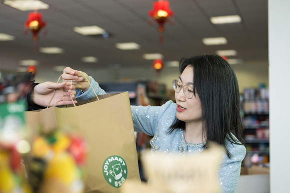 Yaofang (Vicki) Liang hands paper grocery bag to customer. Bag has the logo for JoyMart on it. 