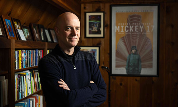Edward Ashton, author of Mickey7, looks at the camera with his arms folded. Behind him is an extensive bookshelf and a framed movie poster for Mickey 17.