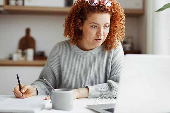 A woman sitting at a table writes on a notepad as she looks at a laptop screen.