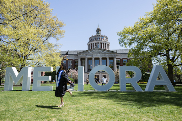 Student in graduation regalia stands in front of large letters spelling Meliora. 