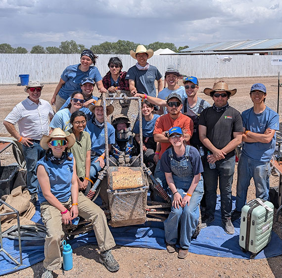 A team of 15 students pose with a muddy baja SAE car on a hot, sunny day in the desert.