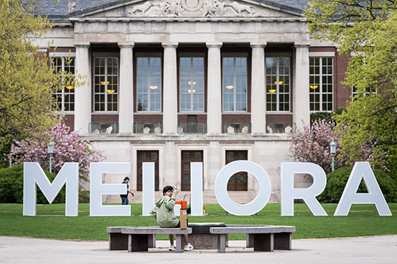 A student takes a selfie in front of the large letters spelling out "MELIORA" on the Eastman Quadrangle.