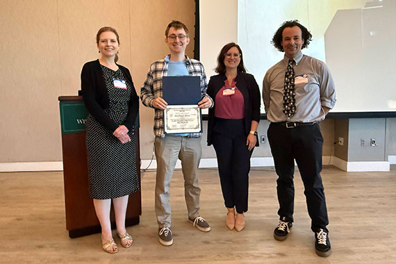 A student accepts a best paper award at a conference while standing next to conference officials and his advisor.