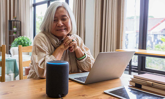A senior woman uses a smart speaker while sitting in front of her laptop in a living room.