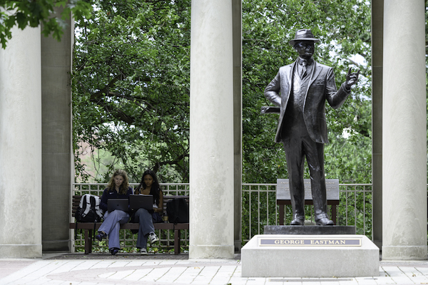 Two students sit on a bench behind the George Eastman Statue on Eastman Quad.
