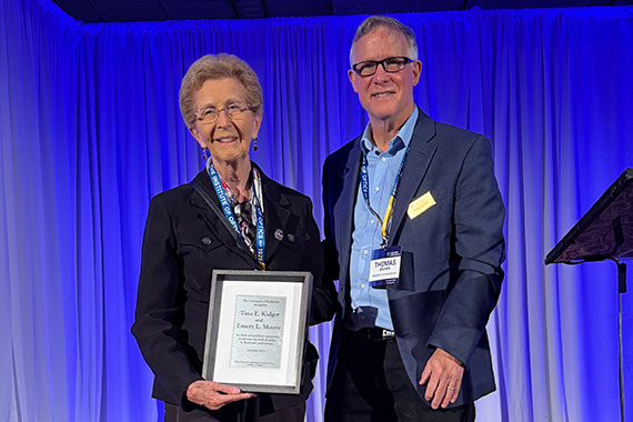 On stage, Tina Kidger holds up a commemorative plaque while standing next to Tom Brown.