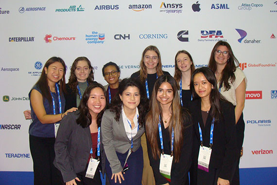 Ten students pose in front of a backdrop featuring the logos of sponsors at the WE25 conference.