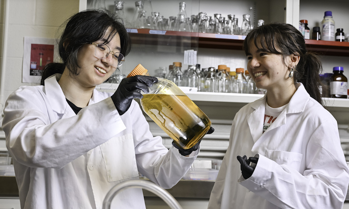 Two students in lab coats examine a bottle of brown liquid.