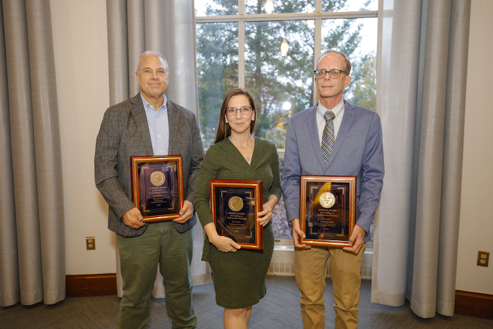Three professors line up in front of window with their awards. 