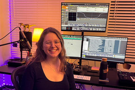 Grace Stensland sits at a computer with three screens displaying audio editing software.