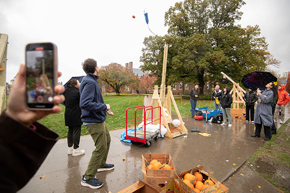 A trebuchet launches a pumpkin in the air on the Wilson Quad on a rainy day as students watch.