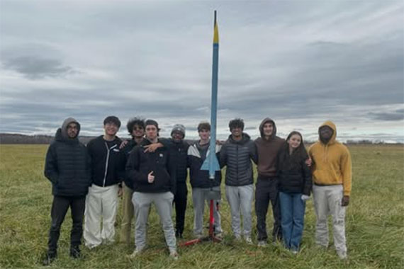 Students stand in front of a large rocket on a cloudy day.