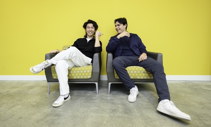 Two students seated next to each other on grey chairs in front of a yellow wall. 