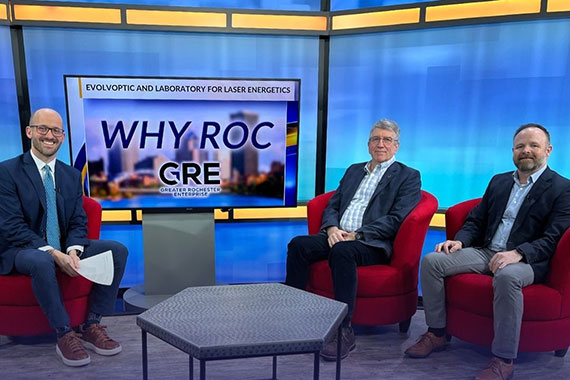 A TV host and two guests sit in a blue studio with a screen that says "Why ROC GRE."