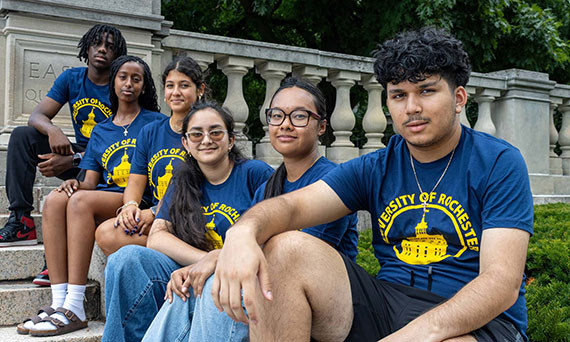 Six high school students wearing University of Rochester T-shirts sit on the stairs leading to the Eastman Quad.