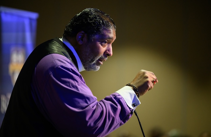 Man in a purple shirt and black vest speaks to a crowd from a microphone at a podium.