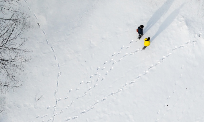 Aerial photo of person in black coat and person in yellow coat walking through the snow. Patterns of footsteps are behind them. 