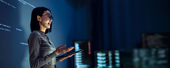 A woman speaking in front of a dark room is lit up by a projector.