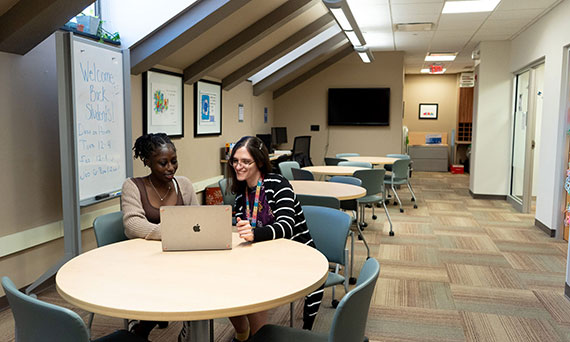 Two women huddle in front of a laptop in a lounge area.