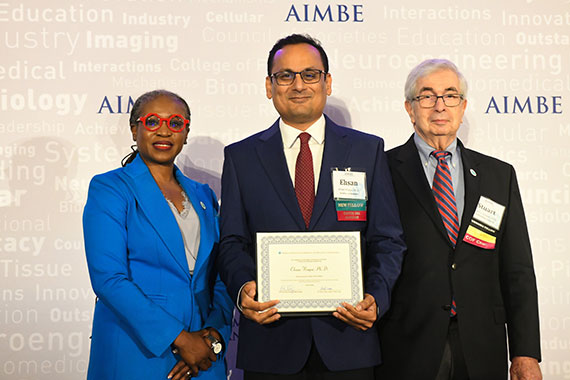 Ehsan Hoque holds a plaque while flanked by two officials in front of an AIMBE backdrop.