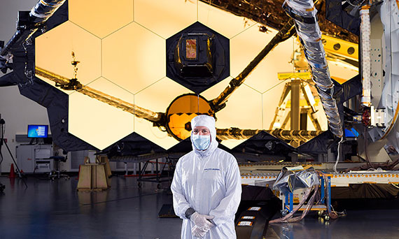 A scientist in a white clean suit stands in front of the golden mirrors of the James Webb Space Telescope.