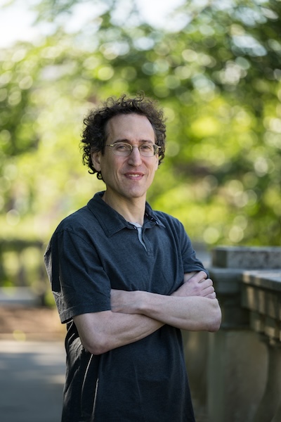 Man in blue shirt standing outside, arms crossed in front of his body.