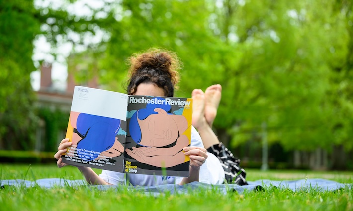 Woman sits upright on blanket reading a copy of a magazine.