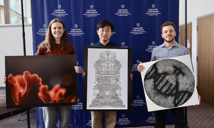 Three students hold their award-winning pieces of art in front of a blue University of Rochester backdrop. 