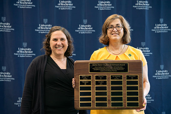 Lisa Altman holds up a plaque for the Dottie Welch Student Enrichment Award while standing next to Wendi Heinzelman in front of a Hajim School backdrop.