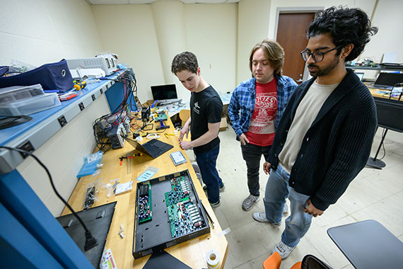 Three students look at circuit boards while they work on their capstone projects in a lab.
