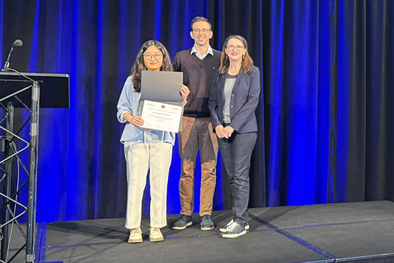 Three people stand on a stage while one holds up a best paper award certificate.