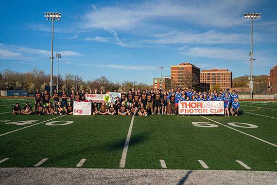 Dozens of students gathered on a football field at the URochester campus hold up signs that say "ThorLabs Photon Cup."