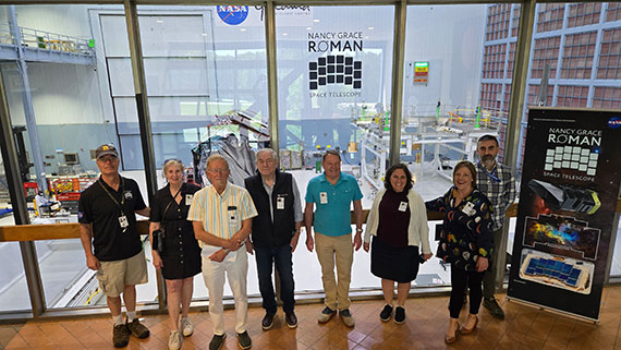 Eight people gather in front of a window at NASA's Goddard Space Flight Center with the Nancy Grace Roman Space Telescope behind them.