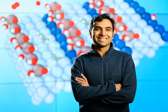 Sid Deshpande stands in front of a screen with a red, white, and blue graphic depciting molecular reactions.