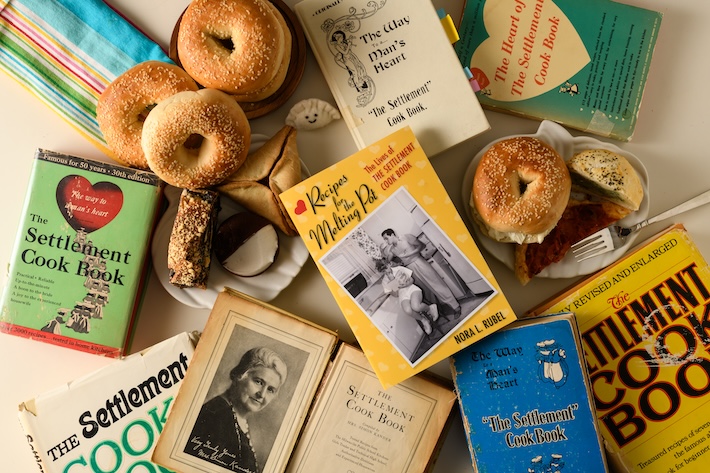 Several cookbooks scattered on a table among plates of bagels and black and white cookies. 
